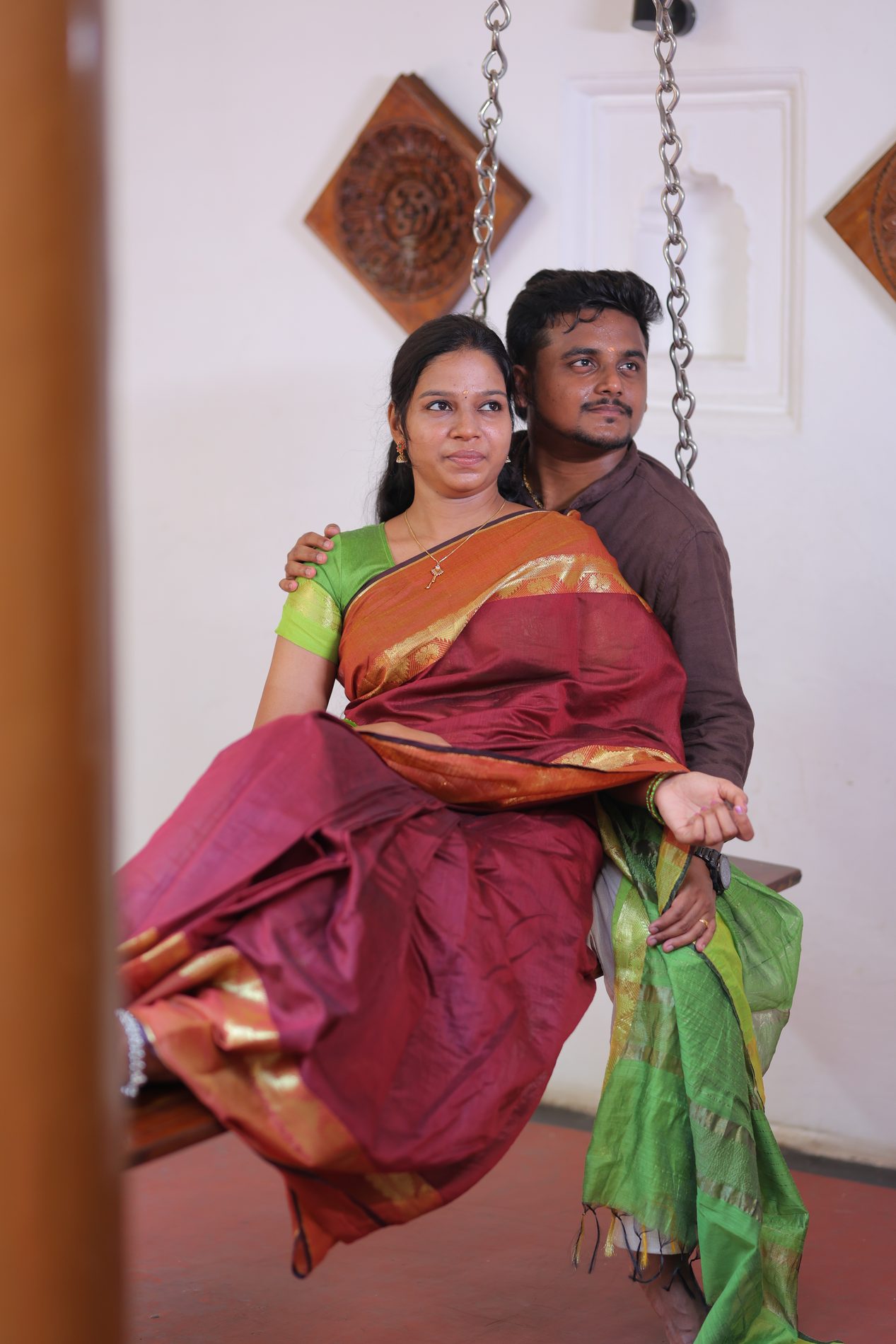 Both together on the oonjal swing, carved wooden wall art behind them